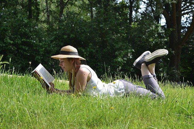 Jeune femme portant un chapeau allongée dans l'herbe et lisant un livre.
