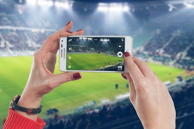 Mains de femme tenant un smartphone devant un stade de football.